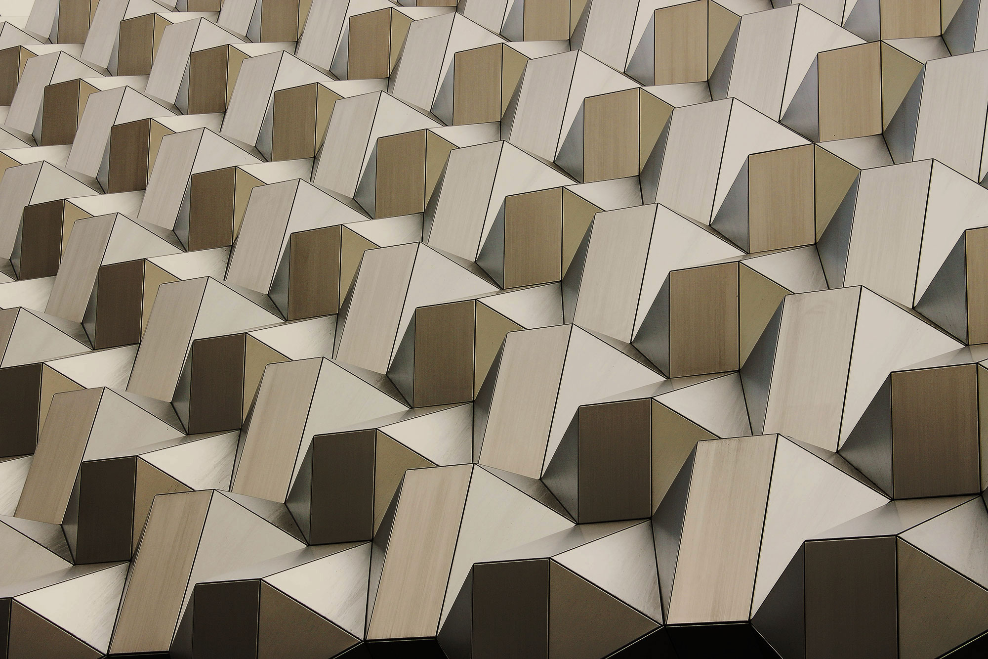 Abstract geometric architectural facade with repeating angular faceted panels in beige, tan, and brown tones, creating a rhythmic modernist pattern